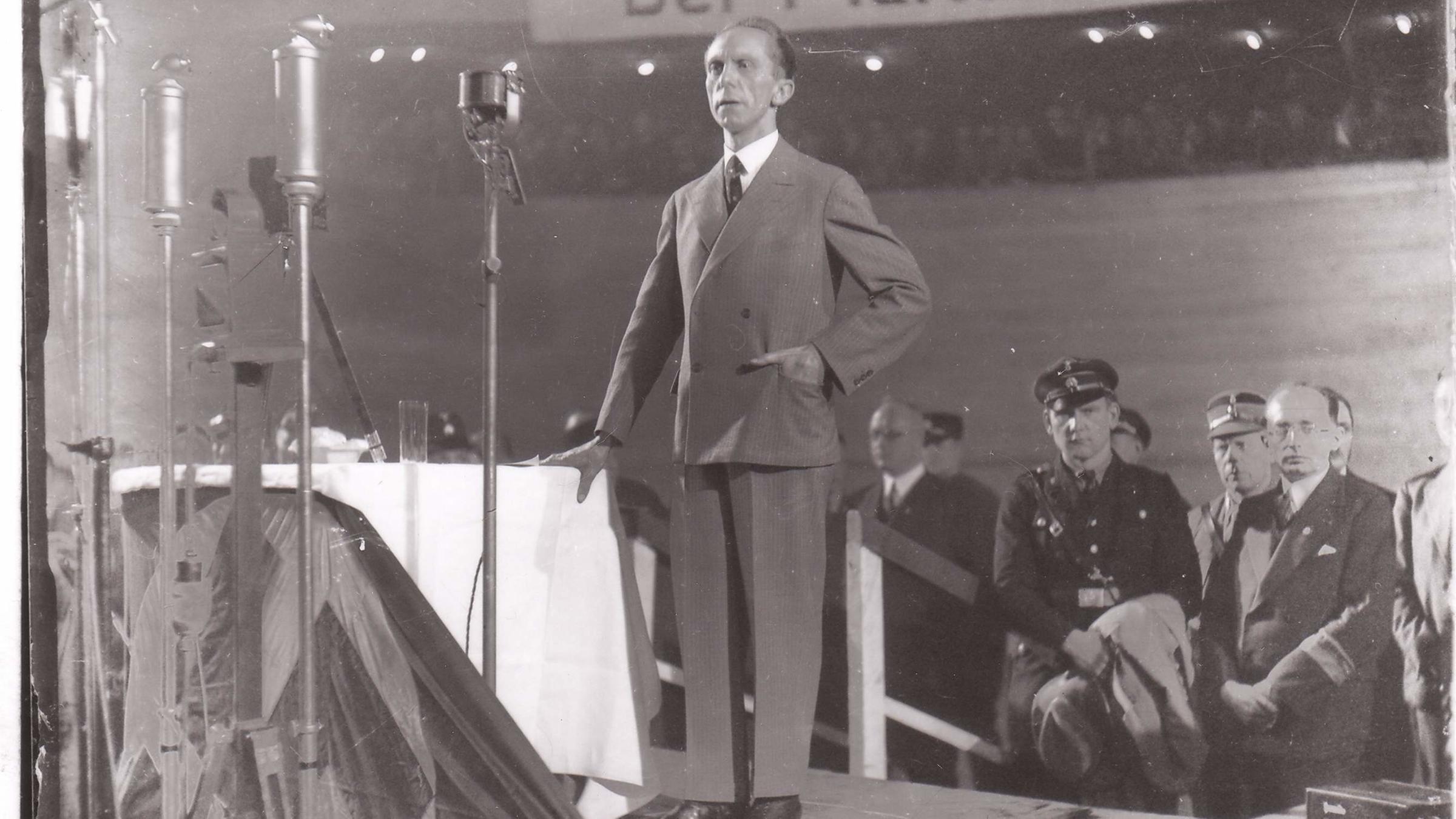 "Propaganda im Krieg - Verblendung der Massen": Propagandaminister Goebbels steht an Tisch auf einer Bühne, mehrere Mikrofone an Ständern, im Hintergrund weitere Männer, oben im Bild ein Spruchband.