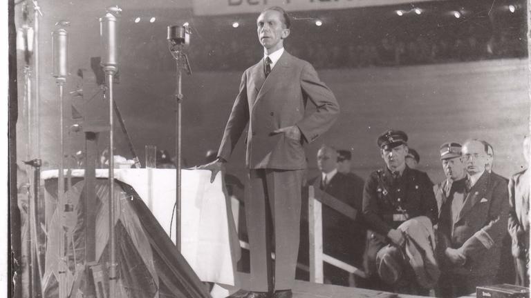 "Propaganda im Krieg - Verblendung der Massen": Propagandaminister Goebbels steht an Tisch auf einer Bühne, mehrere Mikrofone an Ständern, im Hintergrund weitere Männer, oben im Bild ein Spruchband.