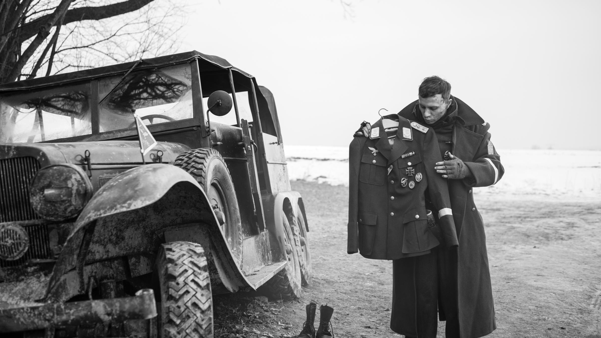"Der Hauptmann": Die linke Bildhälfte wird von einem abgestellten, halb schief stehenden Militärfahrzeug eingenommen. Rechts steht ein junger Soldat, in einen dicken Militär-Wintermantel gehüllt. Er betrachtet eine sauber auf einem Bügel hängende Uniform, die er vor sich in den Händen hält. An der Uniform befindet sich das Eiserne Kreuz nebst anderen Auszeichnungen.