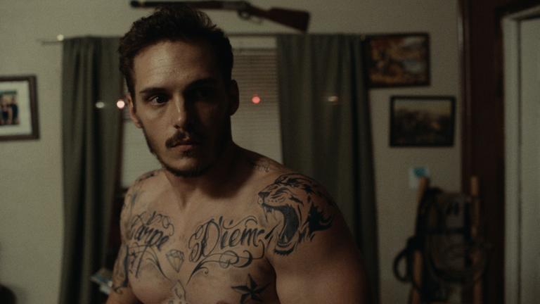 "In der Mitte des Flusses": Großaufnahme Gabriel (Eric Hunter) in einem Zimmer. Er hat verschiedene Tattoos auf dem Oberkörper. Er steht mitten in einem Zimmer und sieht angespannt aus. Über dem Fenster hängt ein Gewehr, an der Wand hängen verschiedene Bilder.