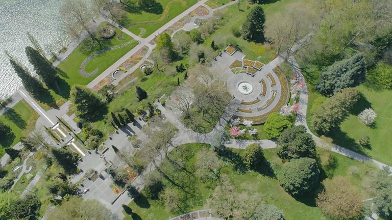 "ZDFzeit: Corona-Land von oben - Natur und Mensch im Lockdown": Die Insel Mainau aus der Luft aufgenommen. Am Rand sieht man den Bodensee. Zu erkennen sind Wiesen, Bäume, leere Spazierwege und Blumenfelder.
