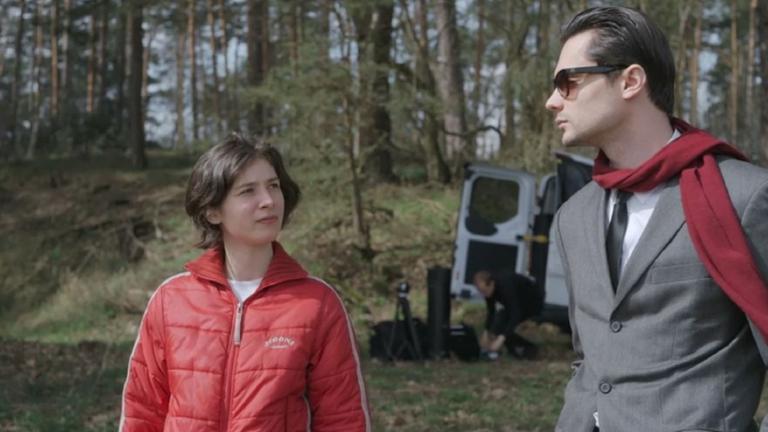 "Which Way to the West": An einem Waldrand, im Hintergrund ein geöffneter Minitrransporter, aus dem ein Mann Filmausrüstung auspackt: Eine junge Frau mit halblangen dunkelblonden Haaren in einem roten Anorak schaut prüfend einen schick angezogenen jungen Mann an, der in grauem Jackett mit weißem Hemd und schwarzer Krawatte, einen roten Wollschal um den Hals, durch seine schwarze Sonnenbrille distanziert auf sie herabblickt.