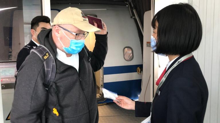 "auslandsjournal - die doku: Inside China - Wie die Pandemie das Land verändert": ZDF-Korrespondent Ulf Röller mit Mundschutz und Reisepass in der Hand steht einer Stewardess mit Mundschutz gegenüber.