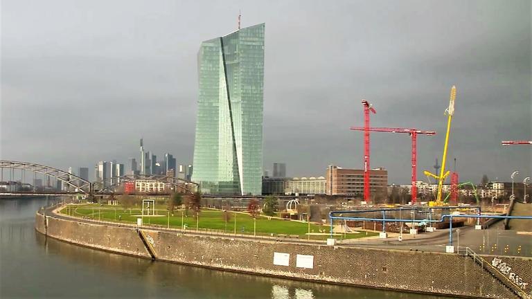 "ZDFzoom: Sparen ade - Wer zahlt den Preis für den Niedrigzins?": Blick auf das hohe Gebäude der EZB in Frankfurt. Im Vordergrund der Main, rechts neben dem EZB-Gebäude befindet sich eine Baustelle mit hohen Kränen. Links hinter dem EZB-Gebäude sieht man die Skyline von Frankfurt.
