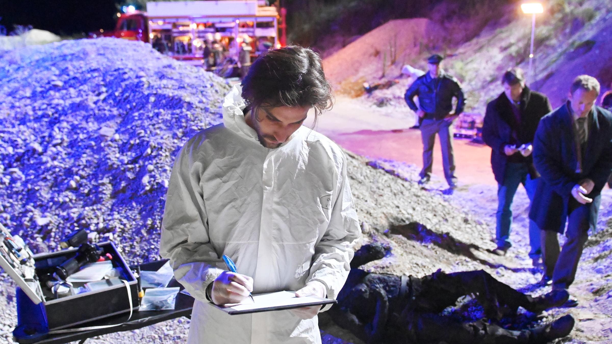 "Aktenzeichen XY-Spezial: Cold Case": Eine Leiche liegt auf einer Lavagrube. Ein Mann im weißen Schutzanzug notiert den Befund. Ein Polizist und zwei weitere Männer stehen seitlich zur Leiche.