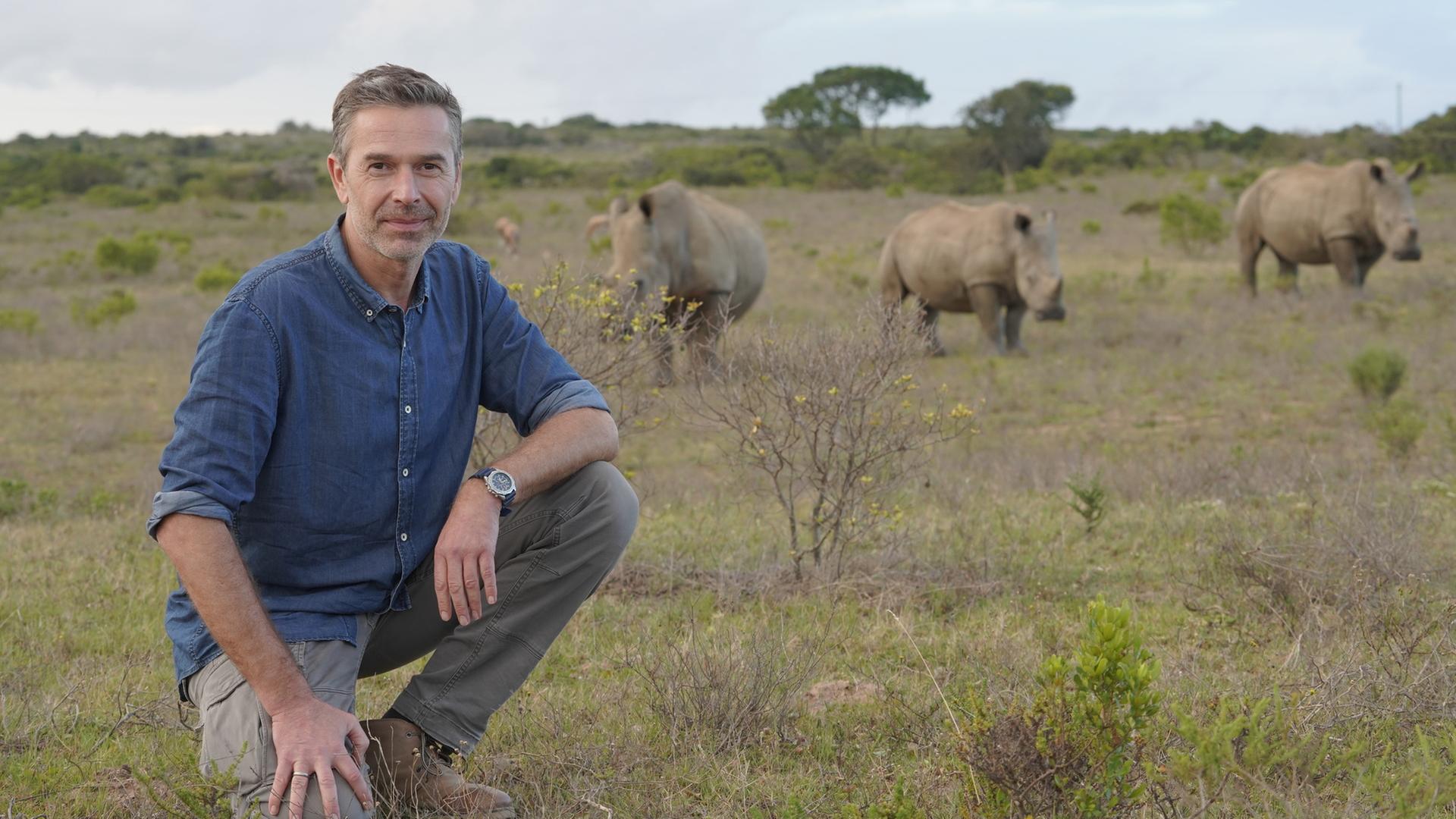 "Terra X - Faszination Erde: Aus Liebe zum Leben: Artenschutz": Dirk Steffens sitzt im Freien und schaut in die Kamera. Im Hintergrund stehen drei Nashörner.