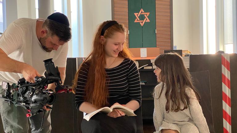 "Hey, Jude! - Jung.Jüdisch.Deutsch.": Meira sitzt in einer Synagoge, rechts neben ihr ein Mädchen, links ein Mann mit Kamera. Im Hintergrund sieht man einen goldenen Davidstern auf dunkelgrünem Grund.