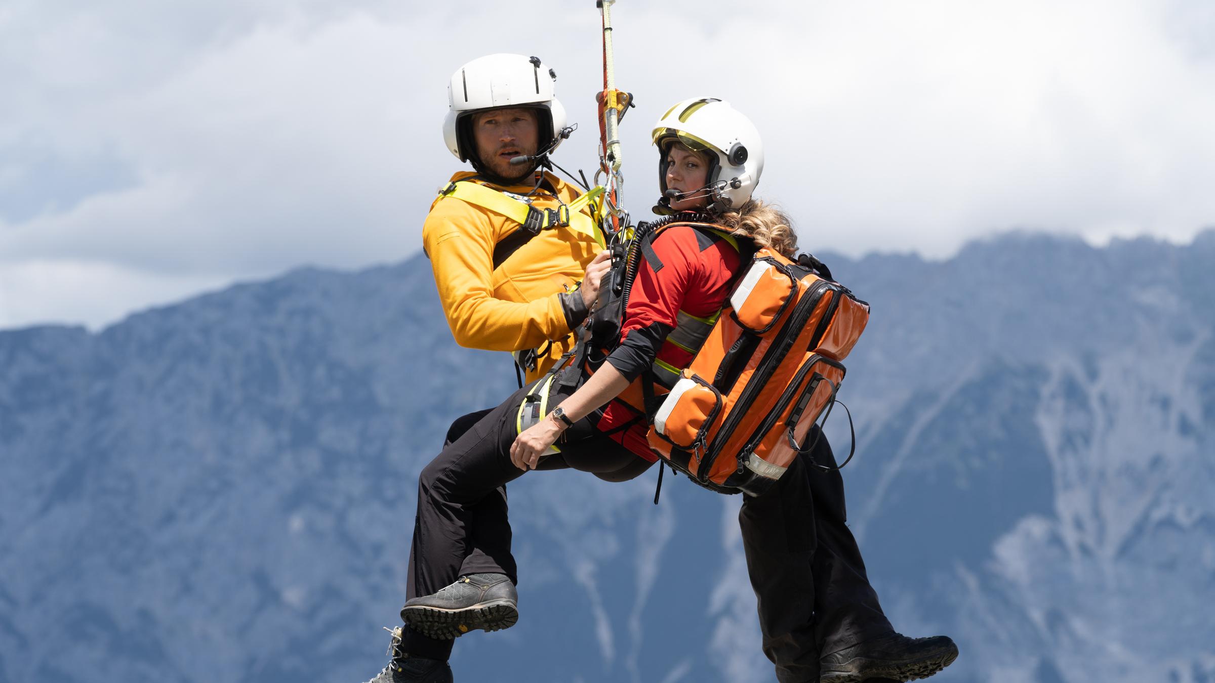 "Die Bergretter: Was wirklich zählt": Die Bergretter Katharina (Luise Bähr) und Markus (Sebastian Ströbel) hängen in ihrer Ausrüstung an einem Seil unter dem Rettungshubschrauber und schweben in der Luft. Im Hintergrund eine Berglandschaft.