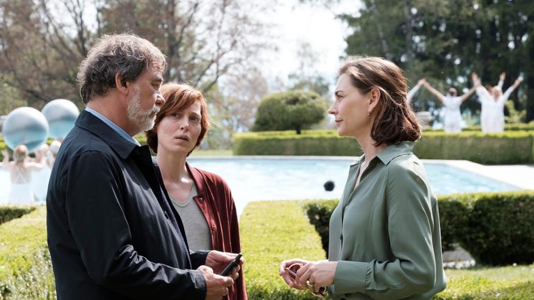 "Die purpurnen Flüsse - Zum Sterben schön": Niémans (Olivier Marchal) und Camille Delaunay (Erika Sainte) stehen vor der Klinikleiterin Vialle (Christiane Paul) und sprechen mit ihr. Im Hintergrund sieht man Hecken und einen großen Pool.