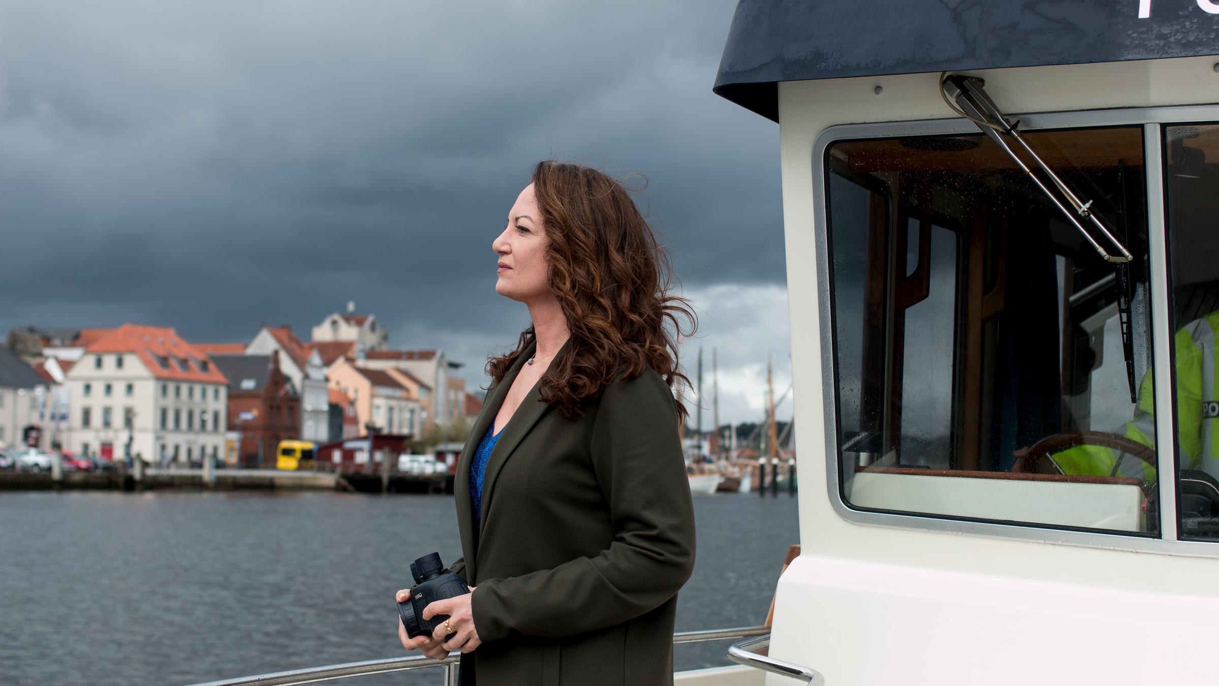 "Unter anderen Umständen – Lügen und Geheimnisse": Jana Winter steht auf dem Deck eines fahrenden Bootes. Sie hält ein Fernglas den Händen. Im Hintergrund sieht man Wasser, einen dunklen, bewölkten Himmel sowie das Ufer und Häuser.