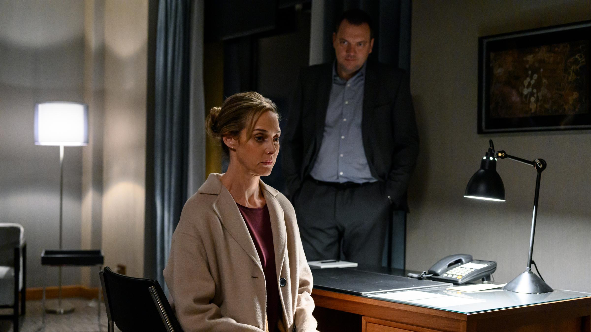 "Das Verhör in der Nacht": Judith (Sophie von Kessel) sitzt auf einem Stuhl an einem Schreibtisch. Auf dem Schreibtisch befindet sich ein Telefon und eine brennende Lampe. Hinter dem Schreibtisch steht Thomas (Charly Hübner), der seine Hände in seinen Hosentaschen hat. Thomas schaut Judith von hinten an, die ins Leere schaut.