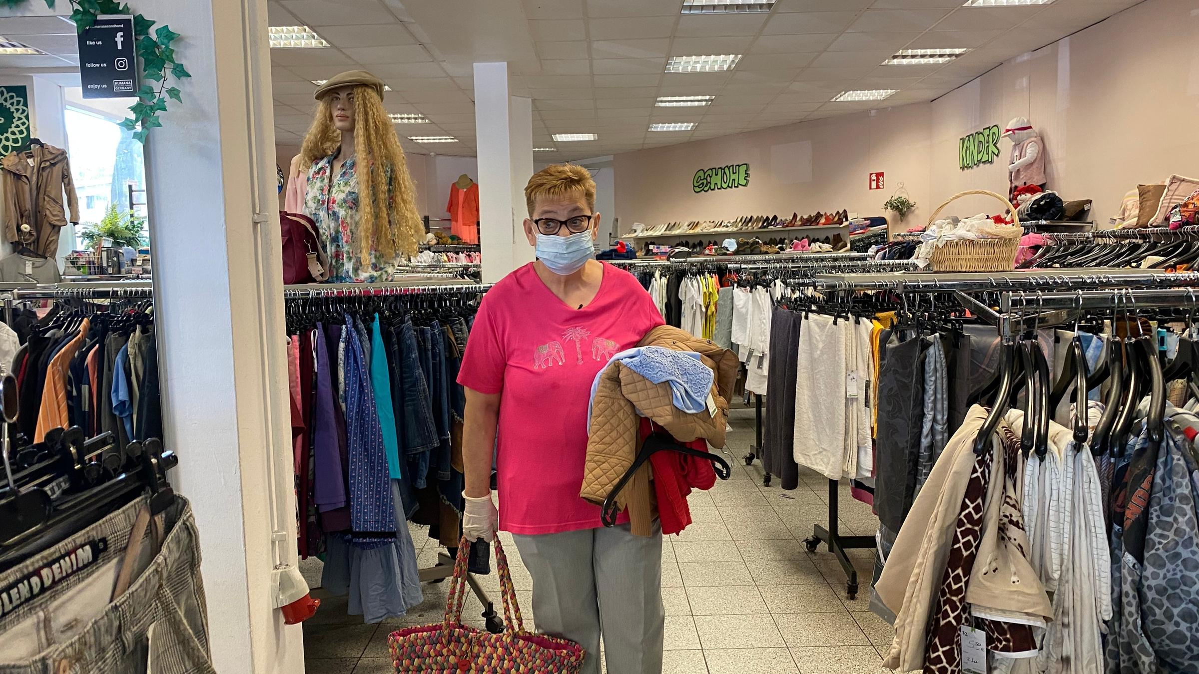 "Dauerstress statt Ruhestand - Reicht die Rente?": Hannelore Janke steht in einem Bekleidungsgeschäft. Sie trägt einen Mundschutz und hält einen Einkaufskorb in der Hand und Kleidung über einem Arm.