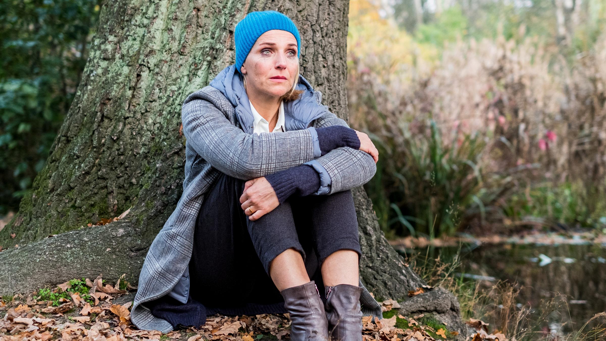 "Fritzie - Der Himmel muss warten - Aus dem Gleichgewicht": Fritzie (Tanja Wedhorn) sitzt auf dem Boden an einen Baum angelehnt. Sie umfasst mit beiden Armen ihre Knie und schaut traurig ins Leere.