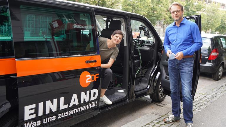 "EINLAND - Was nun zusammenhält": Die Autoren Melanie Haack und Peter Kunz mit dem Kleinbus EINLAND. Melanie Haack sitzt in der geöffneten hinteren Wagentür und schaut heraus, Peter Kunz steht an der geöffneten Beifahrertür. Auf dem Bus steht "EINLAND  - Was uns zusammenhält".