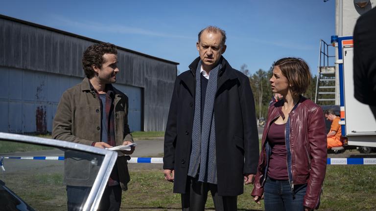 "Nord Nord Mord – Sievers und die schlaflosen Nächte": Carl Sievers (Peter Heinrich Brix), Ina Behrendsen (Julia Brendler) und Hinnerk Feldmann (Oliver Wnuk) stehen innerhalb des abgesperrten Tatorts neben einer geöffneten Autotür und beraten sich. Im Hintergrund sind das blau-weiße Absperrband, ein Hanger und das Heck eines Lieferwagens, auf dessen Trittbrett ein Müllmann (Steffen C. Jürgens) in orangem Overall sitzt, zu sehen.