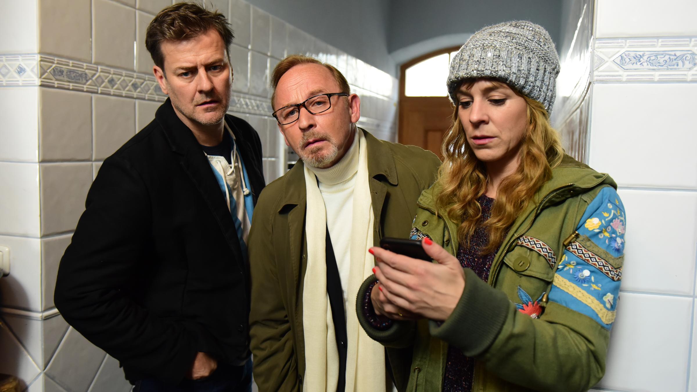 "München Mord - Ausnahmezustand": Harald Neuhauser (Marcus Mittermeier), Ludwig Schaller (Alexander Held) und Angelika Flierl (Bernadette Heerwagen) stehen in einem gekachelten Hausflur. Neuhauser und Schaller blicken interessiert nach vorne, Flierl sieht auf ihr Handy.