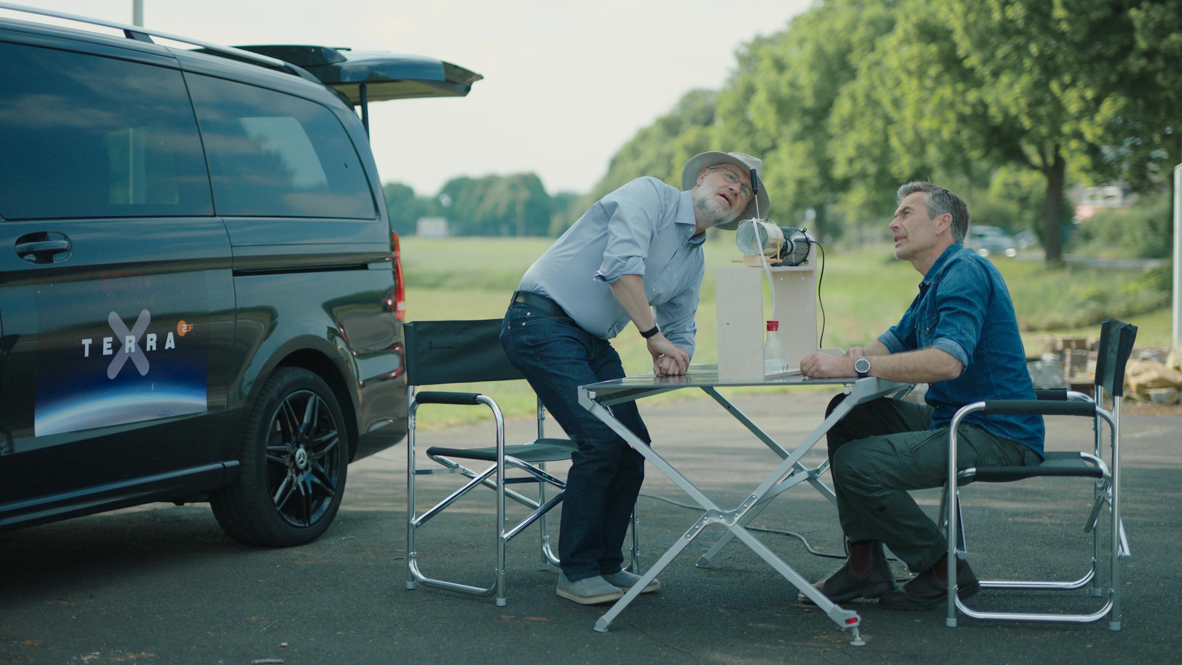 "Ein Fall für Lesch und Steffens - Streiten für die Wahrheit": Harald Lesch und Dirk Steffens sitzen an einem Tisch im Freien. Auf dem Tisch stehen mehrere Gegenstände, auf die die beiden Männer schauen.