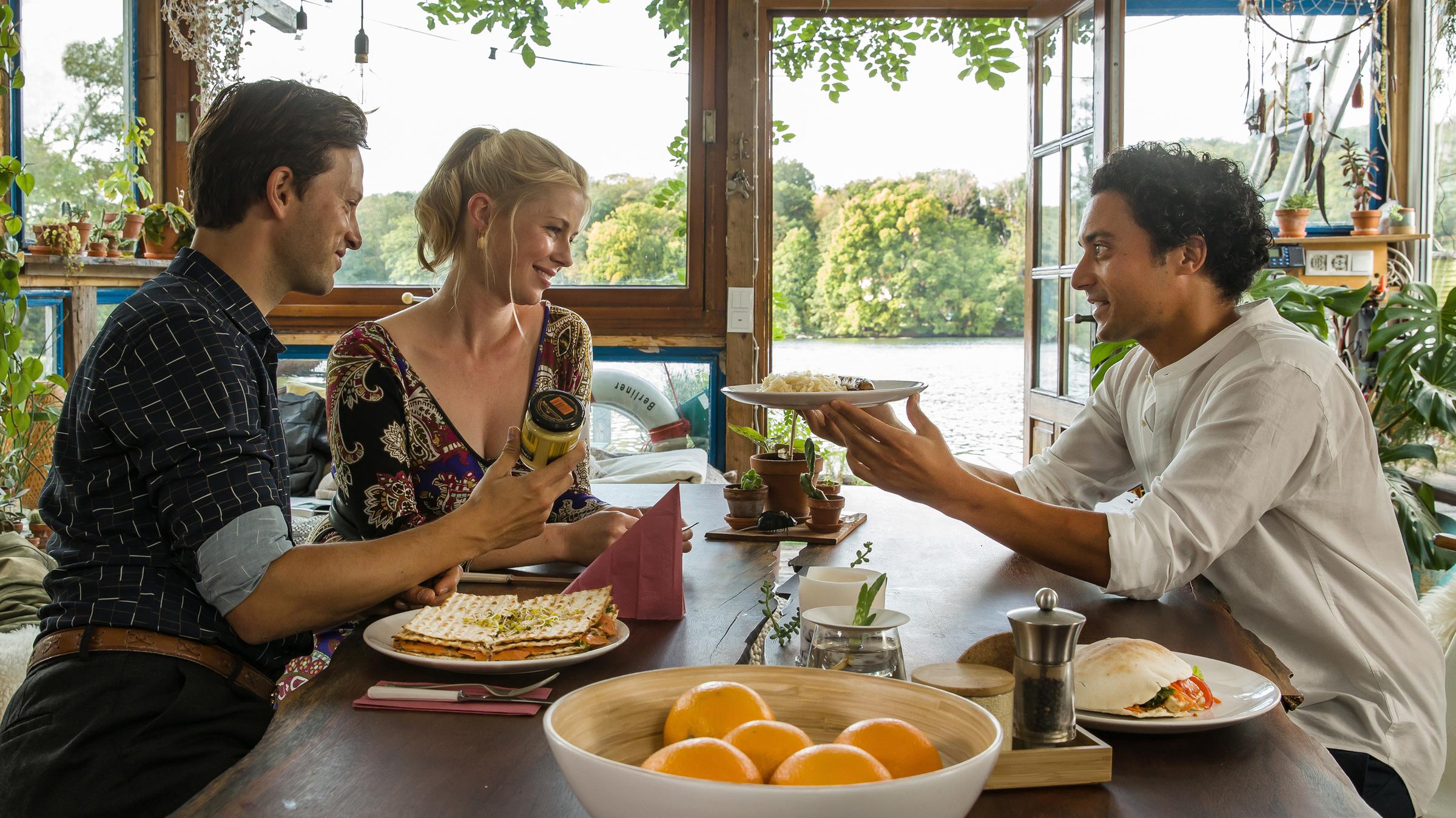 "Matze, Kebab und Sauerkraut": Noah (Franz Dinda), Charlottes (Christine Eixenberger) und Hakim (Omar El-Saeidi) sitzen gemeinsam am Tisch. Hakim hält Charlotte einen Teller hin, im Hintergrund glitzert der See.