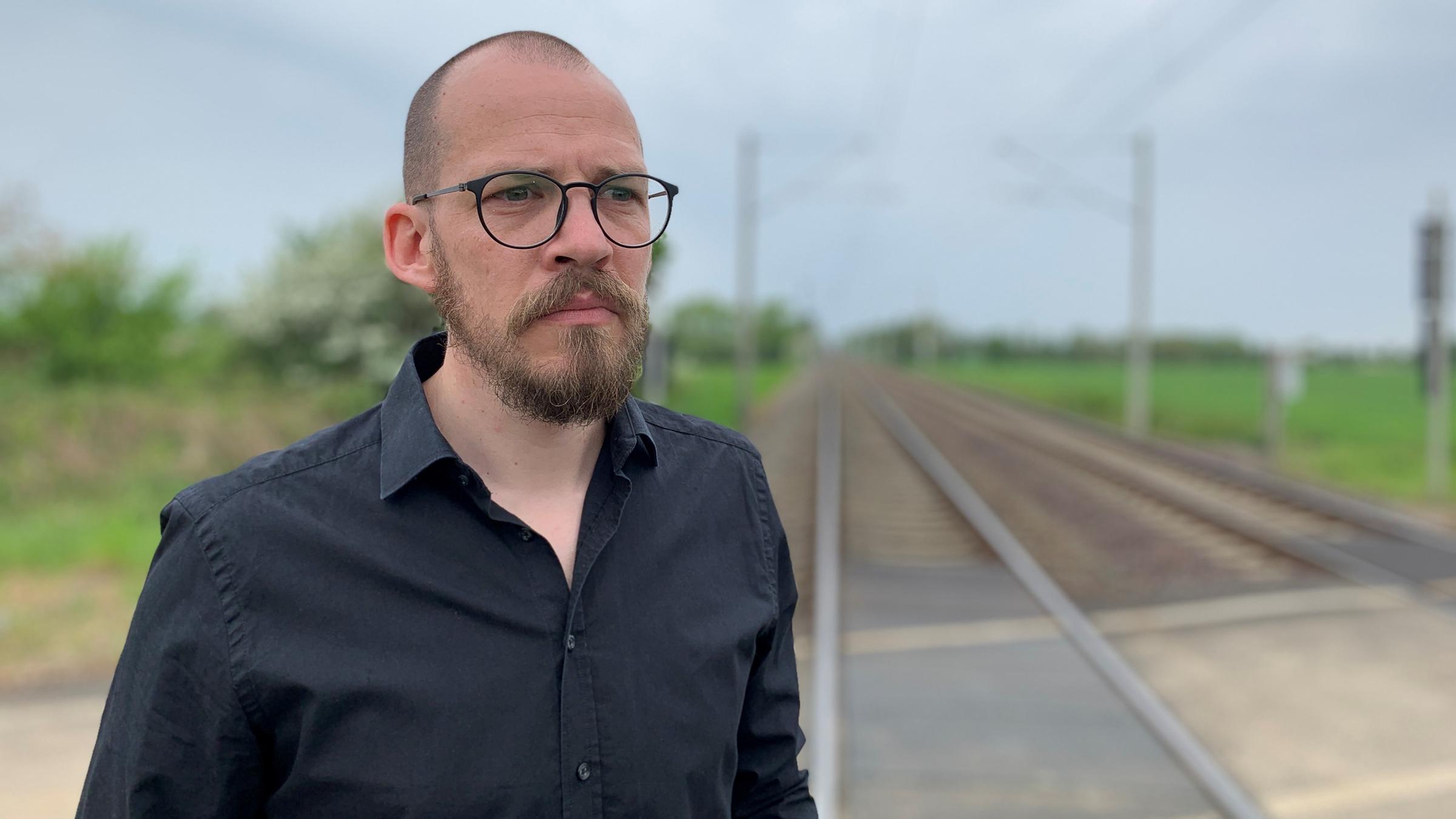 "37° Schatten im Gleis - Wenn Lokführer sich schuldig fühlen": Sören B. steht an einem verlassenen Bahnschienen. Rechts und links von den Gleisen befindet sich das offene Feld.