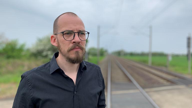 "37° Schatten im Gleis - Wenn Lokführer sich schuldig fühlen": Sören B. steht an einem verlassenen Bahnschienen. Rechts und links von den Gleisen befindet sich das offene Feld.