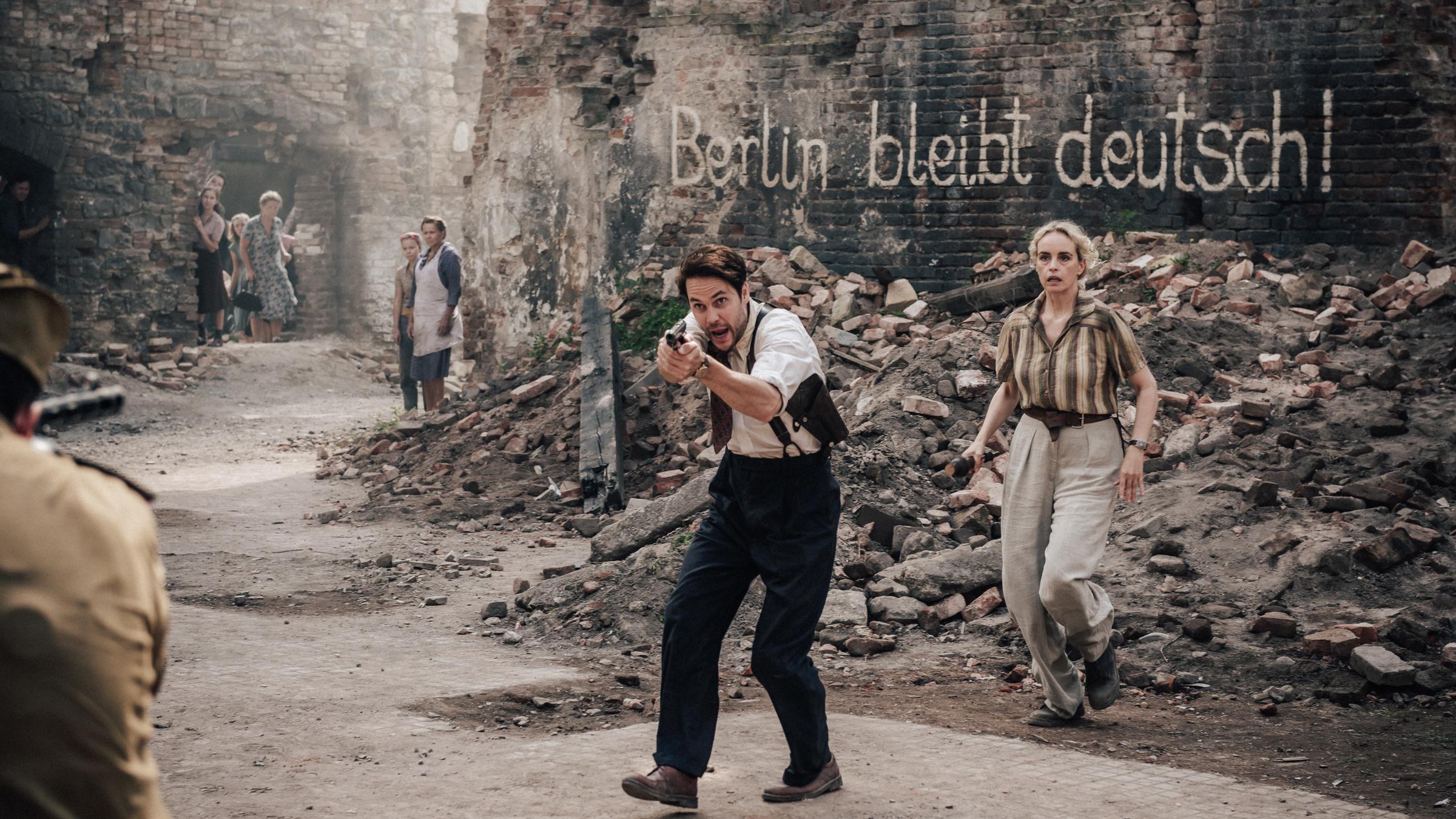 "Schatten der Mörder - Shadowplay (1)": Max McLaughlin (Taylor Kitsch) steht mit gezogener Waffe vor seiner Kollegin Elsie Garten (Nina Hoss), im Hintergrund ein Trümmerhaufen und eine Hauswand mit einem Schriftzug "Berlin bleibt deutsch!".