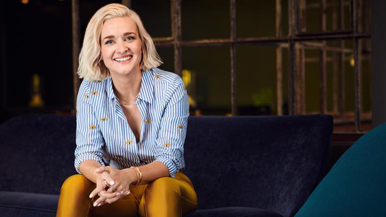 Ariane Alter sitzt mit verschränkten Händen  auf einer Bank im Studio und lächelt in die Kamera.