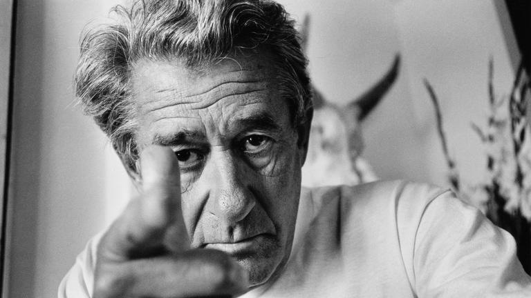 "Helmut Newton - The Bad and the Beautiful": Auf dem schwarz-weiß Bild sieht man den Fotografen Helmut Newton,  er schaut direkt in die Kamera und zeigt mit seinem rechten Zeigefinger direkt auf den Betrachter