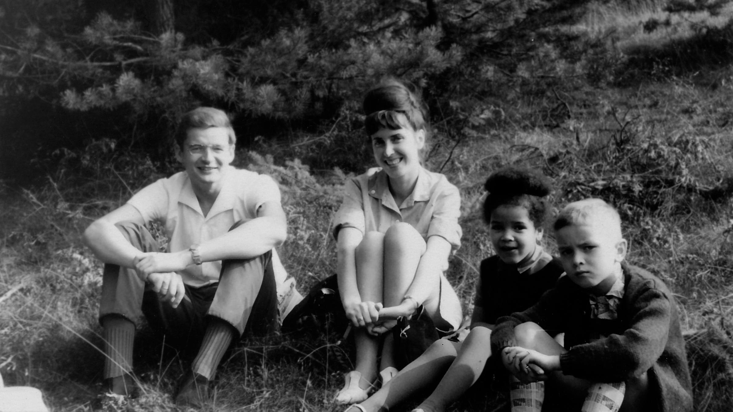 "Becoming Black": Schwarz-Weiß-Aufnahme des Ehepaares Armin und Sigrid Gundermann und den Kindern Ines, einem schwarzen Mädchen und ihrem weißen Bruder Michael. Alle sitzen am Waldrand auf einer Wiese.