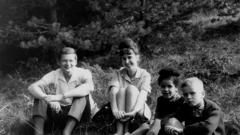 "Becoming Black": Schwarz-Weiß-Aufnahme des Ehepaares Armin und Sigrid Gundermann und den Kindern Ines, einem schwarzen Mädchen und ihrem weißen Bruder Michael. Alle sitzen am Waldrand auf einer Wiese.