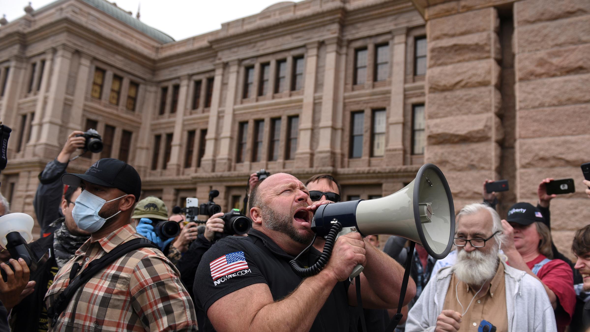 "Labyrinth der Lügen - Trump und die Fake News-Macher": Menschenmenge einer Demonstration vor einem alten Gebäude mit Alex Jones, einem muskulösen Mann im Vordergrund, der in ein Megaphon brüllt.