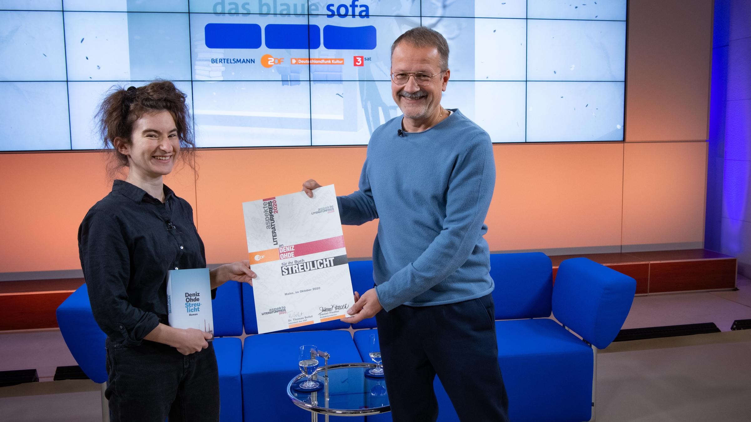 "aspekte Literaturpreis 2020": Deniz Ohde und Daniel Fiedler stehen vor dem Blauen Sofa nebeneinander und halten den Preis in ihren Händen.