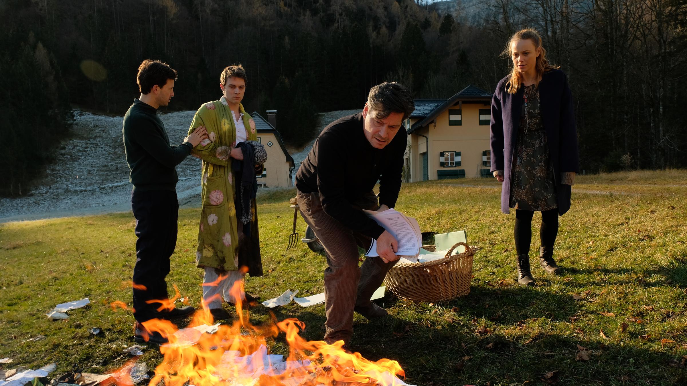 "Das Tal der Mörder": Auf der grünen Wiese vor den Häusern brennt ein Feuer. Die Flammen schlagen hoch und Papier fliegt umher. Drum herum stehen Eva Kufner (Anna Unterberger), sie schaut gespannt auf die Schriftstücke und Anton Gasser (Fritz Karl) wirft weiteres Papier aus einem Weidenkorb in die Flammen. Jakob Gasser (Franz Dinda) hält Florian Gasser (Gerrit Klein) zurück. Florian trägt einen Bademantel.