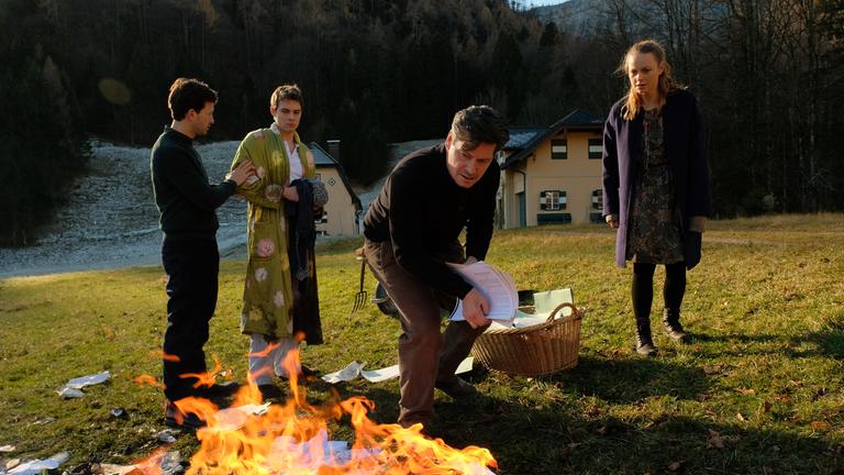 "Das Tal der Mörder": Auf der grünen Wiese vor den Häusern brennt ein Feuer. Die Flammen schlagen hoch und Papier fliegt umher. Drum herum stehen Eva Kufner (Anna Unterberger), sie schaut gespannt auf die Schriftstücke und Anton Gasser (Fritz Karl) wirft weiteres Papier aus einem Weidenkorb in die Flammen. Jakob Gasser (Franz Dinda) hält Florian Gasser (Gerrit Klein) zurück. Florian trägt einen Bademantel.