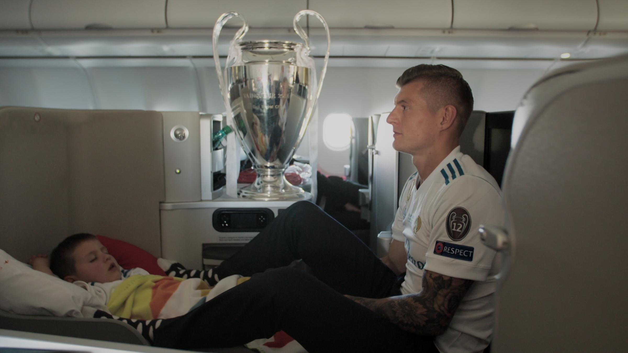 "KROOS": Toni Kroos sitzt in der Firstclass im Flugzeug auf dem ausgeklappten Sitz, auf dem sein Sohn schläft. Der Champions-League-Pokal steht auf einer Ablage.
