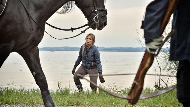 "Die Toten vom Bodensee - Blutritt": Am linken Bildrand ist ein Pferd zu erkennen. Im Anschnitt ist eine Person zu erkennen, die mit Handschuhen eine Armbrust nach unten hält. Im Hintergrund bewegt sich Marlene Stöhr (Stefanie von Poser) auf das Pferd und die Person zu. Hinter ihr befindet sich der Bodensee. Die Frau trägt Reitkleidung. es ist zu erkennen, dass etwas aus ihrem Rücken ragt. Aus ihrem Mund läuft Blut.