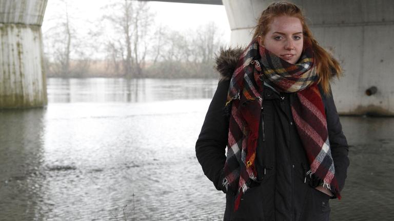 "37°: Ich bin noch da - Suizidgedanken junger Menschen": Junge Frau mit roten langen Haaren, teilweise mit einer Haarklammer zurückgesteckt, steht in Anorak und Schal in einer Fluss-Landschaft.