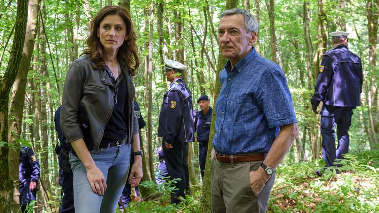 "In Wahrheit - Jagdfieber": Judith Mohn (Christina Hecke) und Markus Zerner (Rudolf Kowalski) stehen im Wald und besprechen sich. Im Hintergrund suchen mehrere Polizisten nach Hinweisen auf den Täter und sichern Spuren.