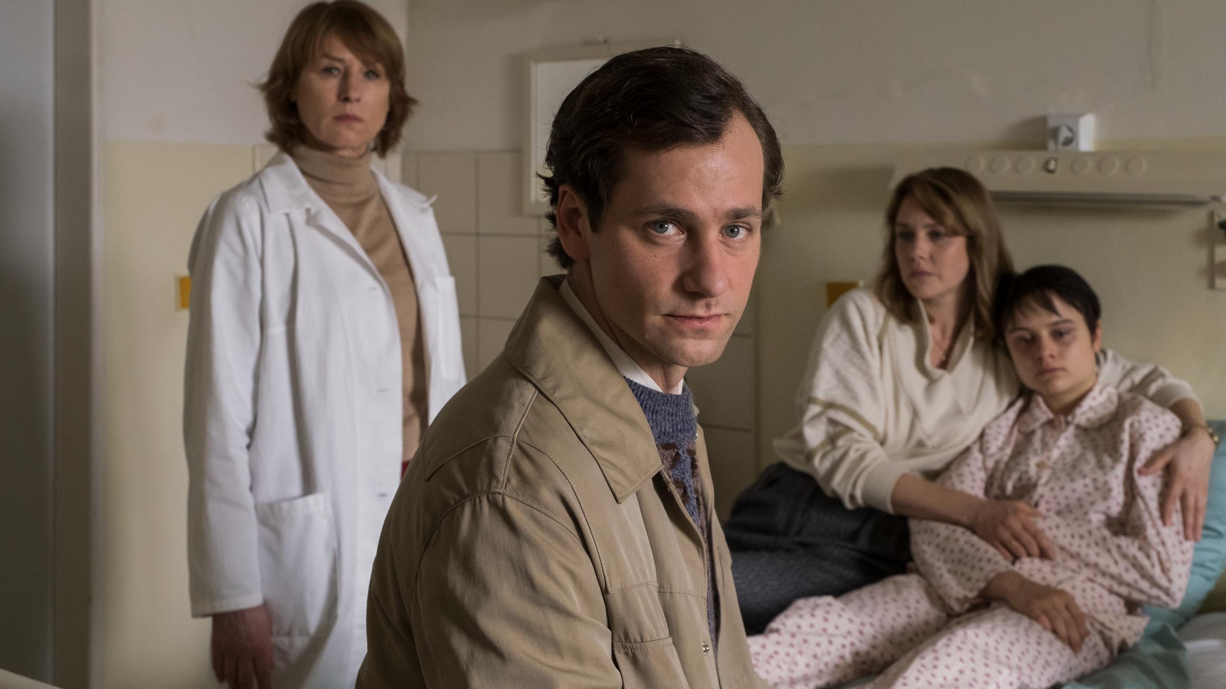 "Kranke Geschäfte": Armin Glaser sitzt im Vordergrund auf dem Krankenbett seiner Tochter Kati Glaser (Lena Urzendowsky) und schaut direkt in  die Kamera. Er greift mit einer Hand hinter sich an Katis Bein, welche im Schlafanzug auf ihrem Bett sitzt, während ihre Mutter Marie Glaser (Felicitas Woll) sie fest im Arm hält. Neben dem Bett im Hintergrund steht Dr. Sigurd (Corinna Harfouch), die mit starrem Blick in Armins Richtung schaut.