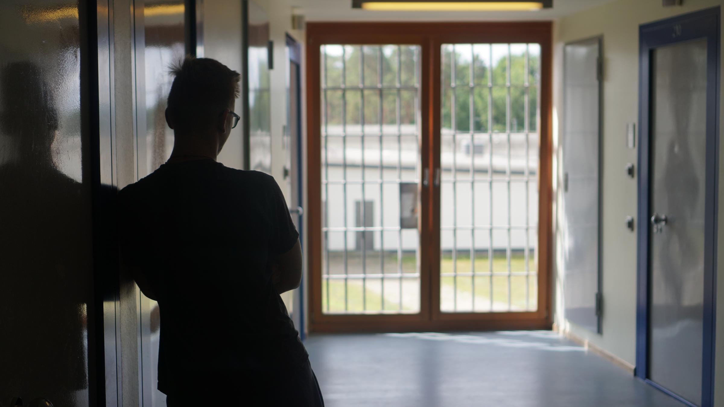 "Wenn Kinder Täter werden": Man sieht von hinten einen Jungen, der auf einen Flur an die linke Wand gelehnt steht. Hinten eine sonnenbeschiene, verglaste Eingangstür. Draußen sieht man ein weißes einstöckiges Gebäude.