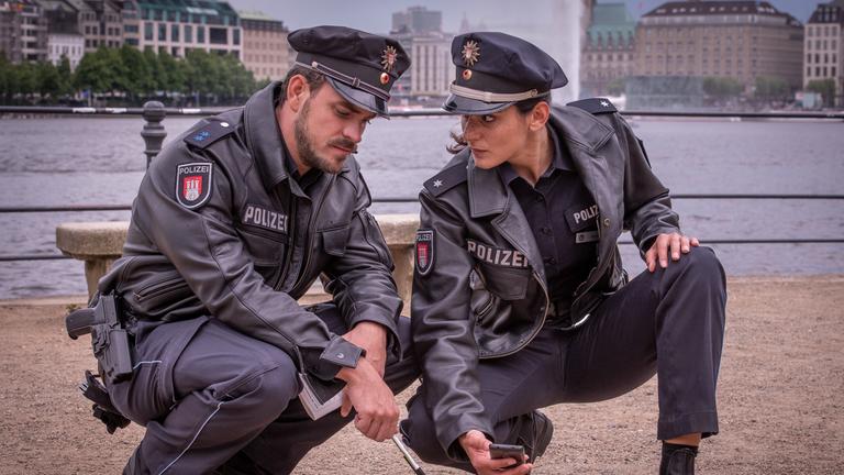 "Notruf Hafenkante - Clangirl": Der Polizist Kris Freiberg (Marc Barthel) und seine Kollegin Pinar Aslan (Aybi Era) sitzen nebeneinander in der Hocke. Pinar hält in ihrer rechten Hand ihr Handy, mit dem sie etwas, das auf dem Boden vor ihnen liegt fotografiert. Sie sieht ihren Kollegen an. Kris hält in der linken Hand ein Notizbuch, in der rechten einen Stift, mit dem er auf das Objekt zeigt.