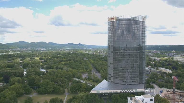 "30 Jahre Deutsche Einheit: Boomtown Bonn - Neuer Glanz in alter Hauptstadt": Blick auf Bonn. Im Vordergrund sieht man den ovalen, gläsernen Turm des Posttowers, in der Mitte die von vielen Bäumen gesäumte Stadt, im Hintergrund die Höhenzüge des Siebengebirges.