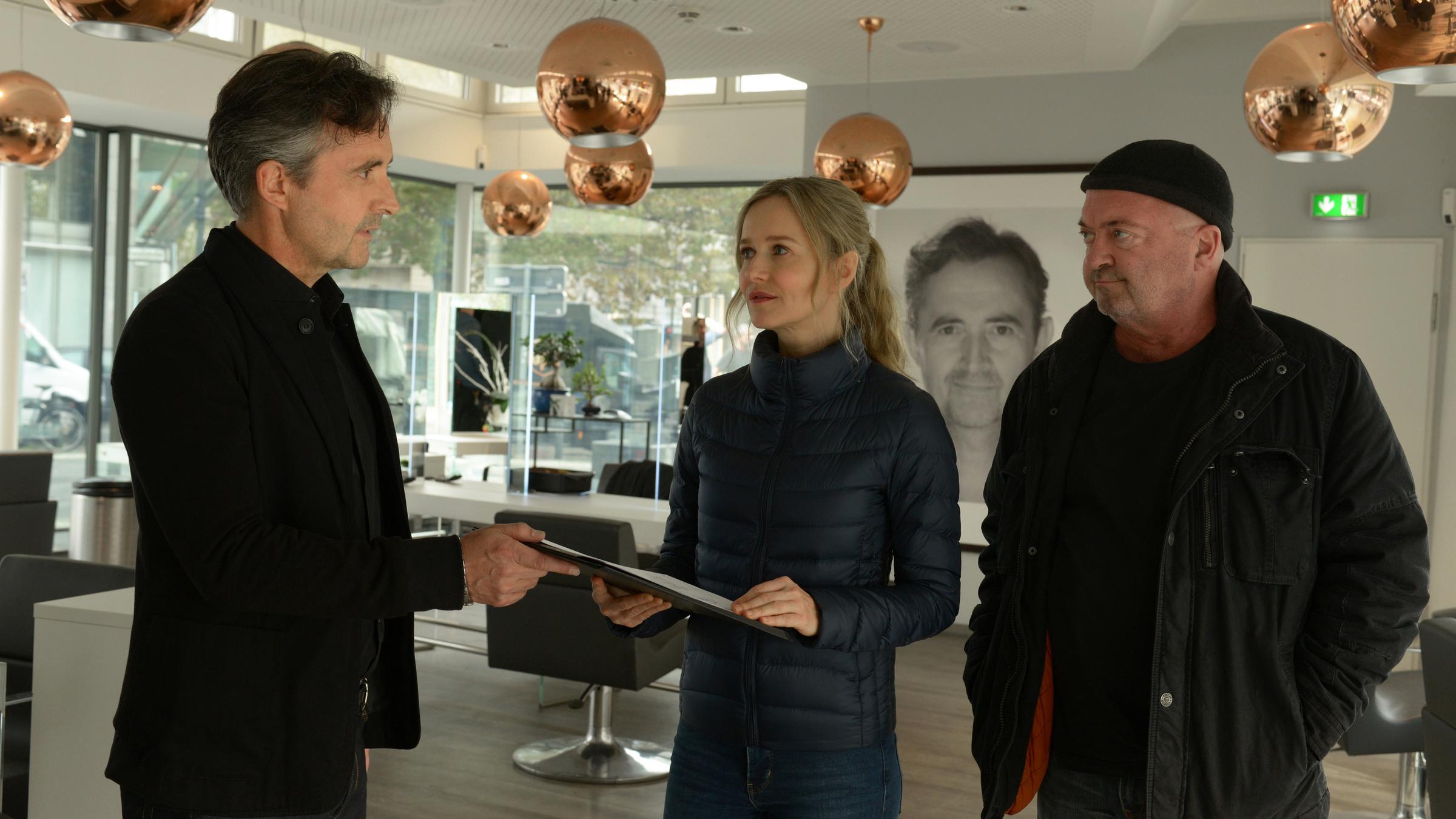 "Ein starkes Team - Scharfe Schnitte": Benno Rohde (Harald Schrott) überreicht Linett Wachow (Stefanie Stappenbeck) Papiere. Otto Garber (Florian Martens) steht daneben, beide Hände sind in seinen Jackentaschen vergraben, während er skeptisch zu Rohde schaut.