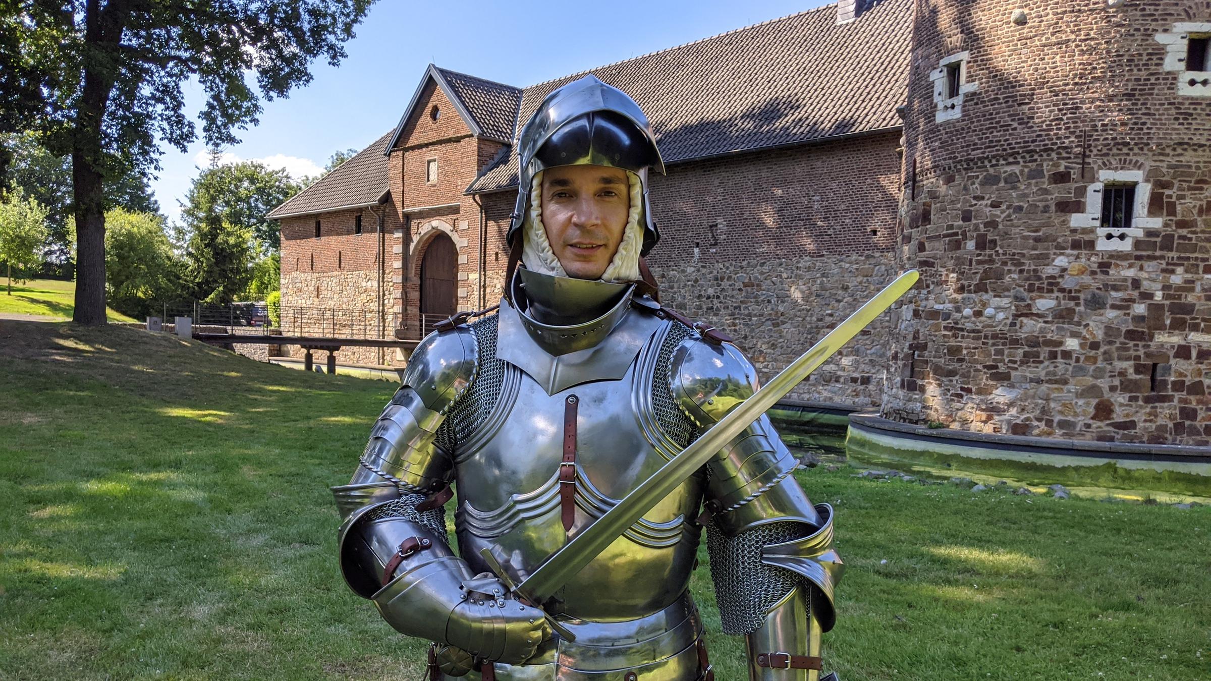 "PUR+- Ritter - Kampf mit der Rüstung": Moderator Eric Mayer trägt eine silbern glänzende Ritterrüstung. In der rechten Hand hält er ein Schwert. Eric steht vor einer mittelalterlichen Wasserburg.