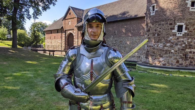 "PUR+- Ritter - Kampf mit der Rüstung": Moderator Eric Mayer trägt eine silbern glänzende Ritterrüstung. In der rechten Hand hält er ein Schwert. Eric steht vor einer mittelalterlichen Wasserburg.