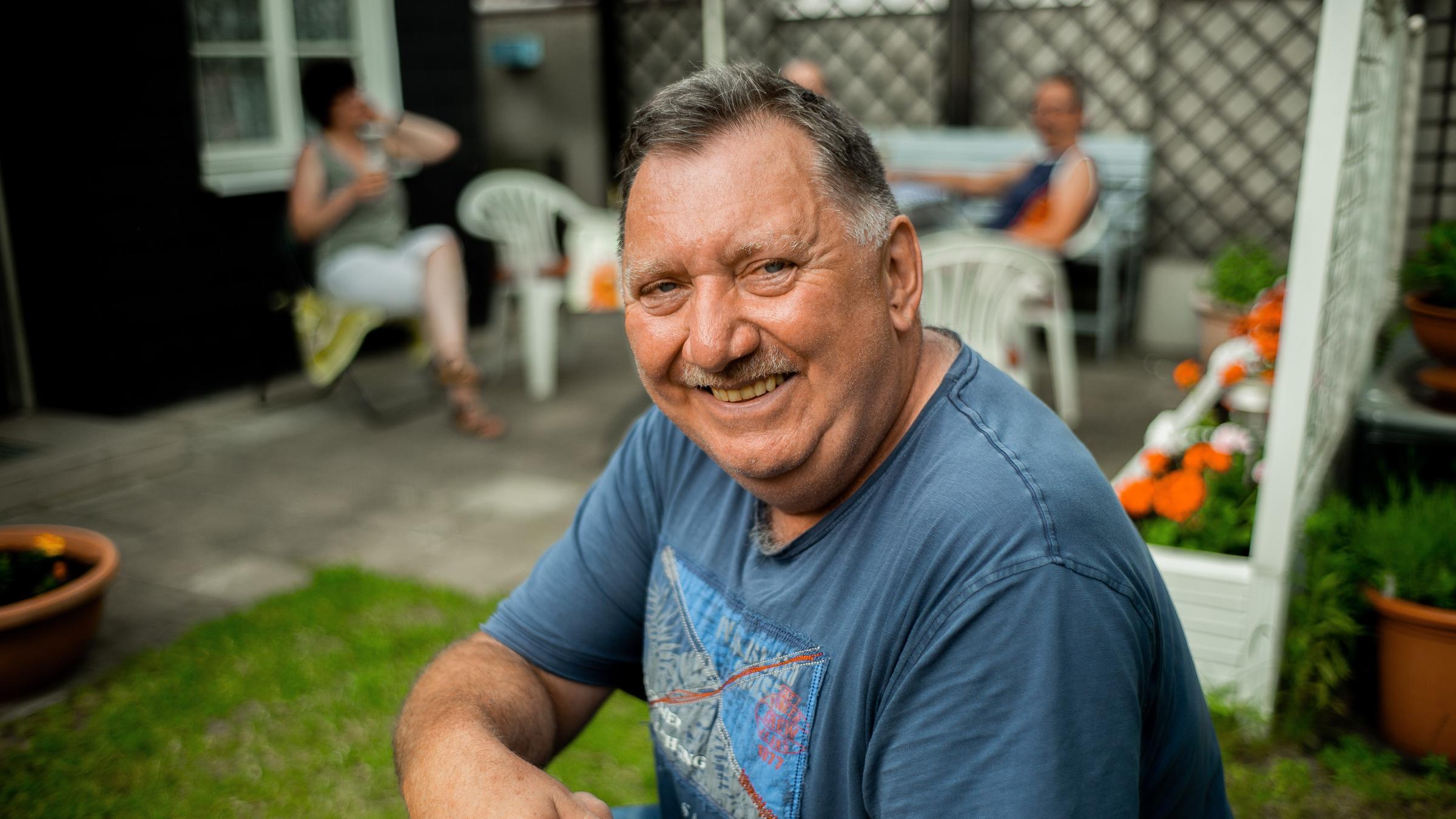 "Wir 80 Millionen. Was Deutschland vereint.": Ein Mann lächelt in die Kamera; im Hintergrund sitzen zwei Menschen auf einer Terrasse.