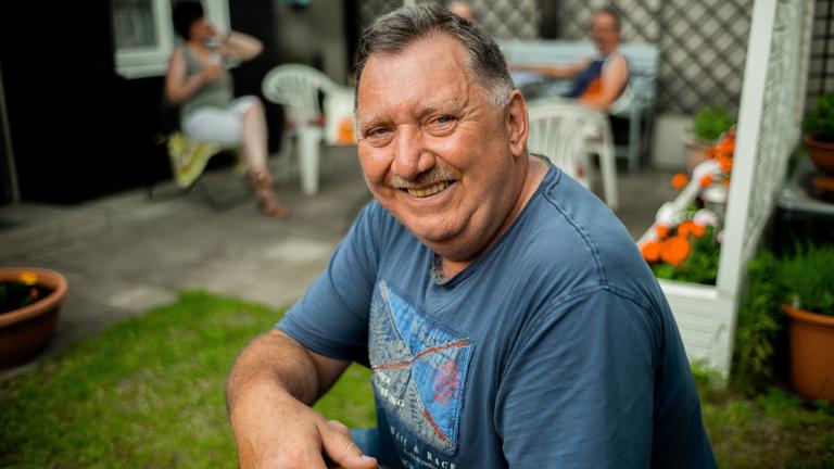 "Wir 80 Millionen. Was Deutschland vereint.": Ein Mann lächelt in die Kamera; im Hintergrund sitzen zwei Menschen auf einer Terrasse.