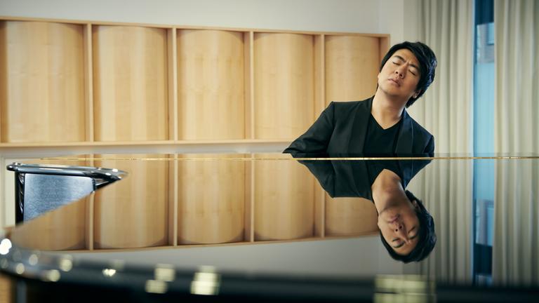 "Lang Langs Goldberg-Variationen": Blick von vorne auf den Pianisten Lang Lang, der mit geschlossenen Augen an einem schwarz-glänzenden Flügel sitzt und spielt. Sein Gesicht spiegelt sich in der Oberfläche des Flügels. Er trägt einen schwarzen Anzug und ein schwarzes T-Shirt.