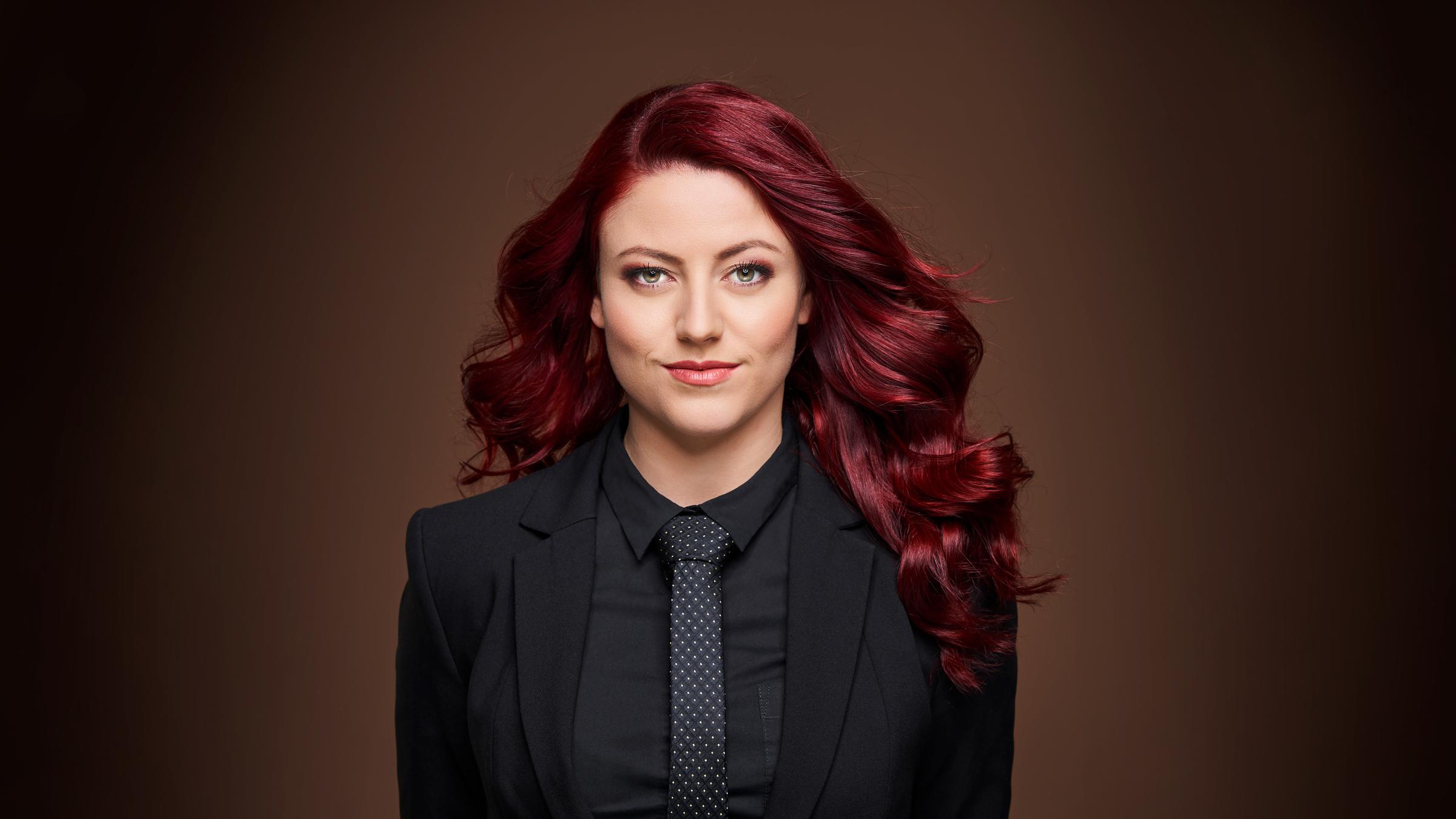 "Tahnee: Vulvarine": Portrait von Tahnee. Sie trägt ihre roten Haare leicht wellig und offen. Gekleidet ist sie mit einem schwarzen Hemd, einer schwarzen Krawatte mit kleinen weißen Punkten und einem schwarzen Blazer.