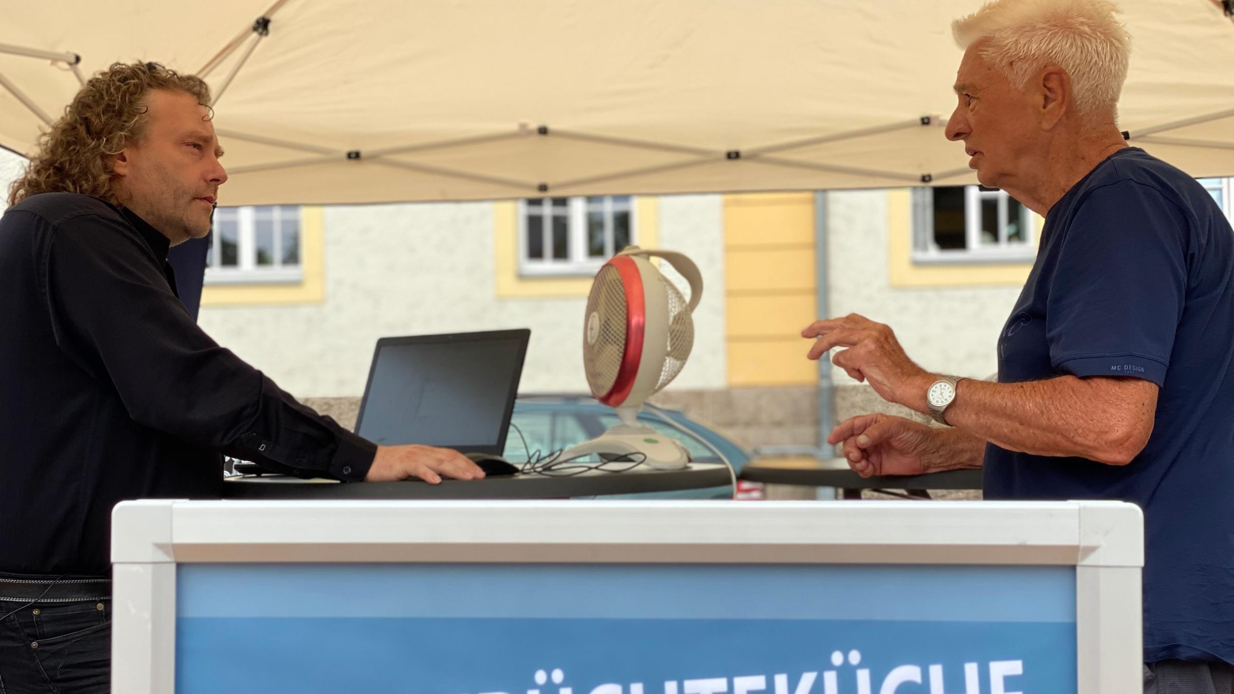 "ZDFzoom: Feindbild Politiker - Sorge um die Demokratie": Oberbürgermeister Torsten Pötzsch steht unter einem Sonnenbaldachin an einem runden schwarzen Tisch, auf dem ein Ventilator und ein Laptop zu sehen sind. Er unterhält sich mit einem Mann, der ihm gegenüber steht. Vor ihnen sieht man ein blaues Plakat, auf dem in weißer Schrift steht: OBs Gerüchteküche.