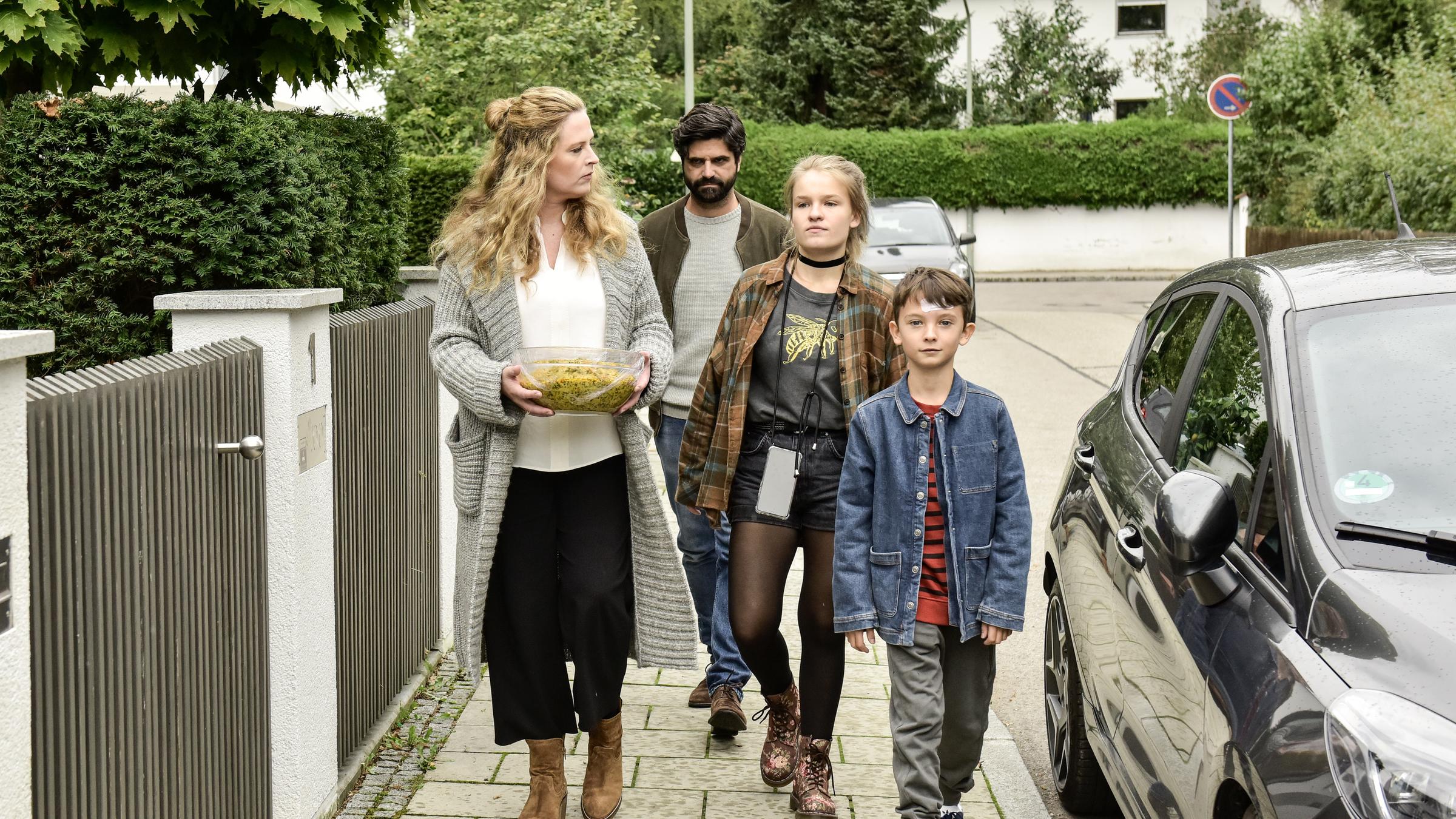 "Zum Glück zurück": Anne (Diana Amft), Nils (Maximilian Grill), Mascha (Luisa Römer) und Lenny (Fabian Ziems) gehen auf einem Bürgersteig entlang. Links im Bild befindet sich ein Gartenzaun, rechts ein parkendes Auto. Anne trägt einen Salat und schaut Mascha an. Nils schaut grimmig.